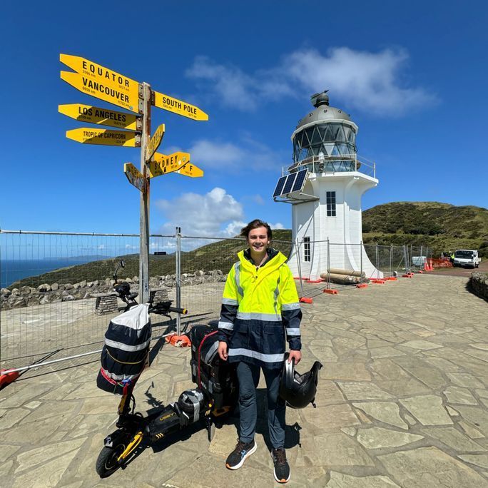 Photo of a man posing before a lighthouse while standing next to his e-scooter.