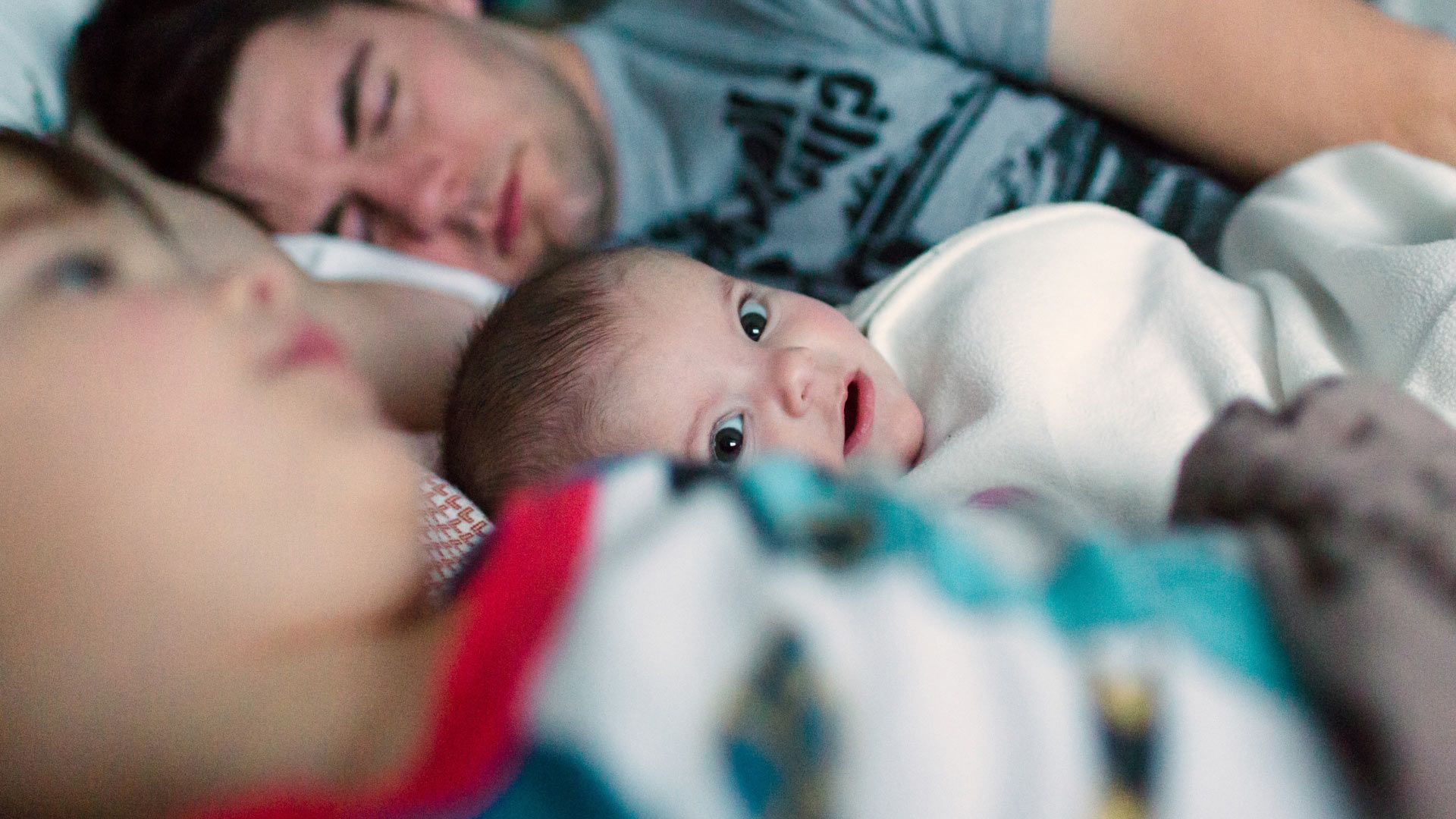 Photo of infant looking to camera between his sleeping father and a sibling